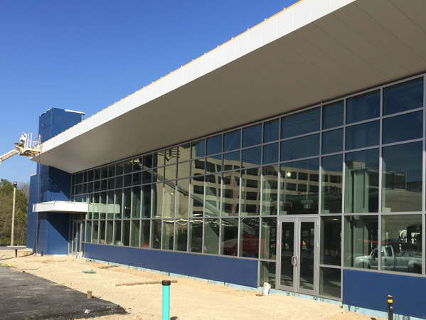 Exterior view of a modern pre-engineered steel building with large glass windows and a blue and white color scheme. The building appears to be under construction, with some construction materials and equipment visible in the foreground.